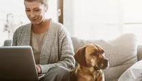 Woman using a laptop whilst sat on a sofa with a dog